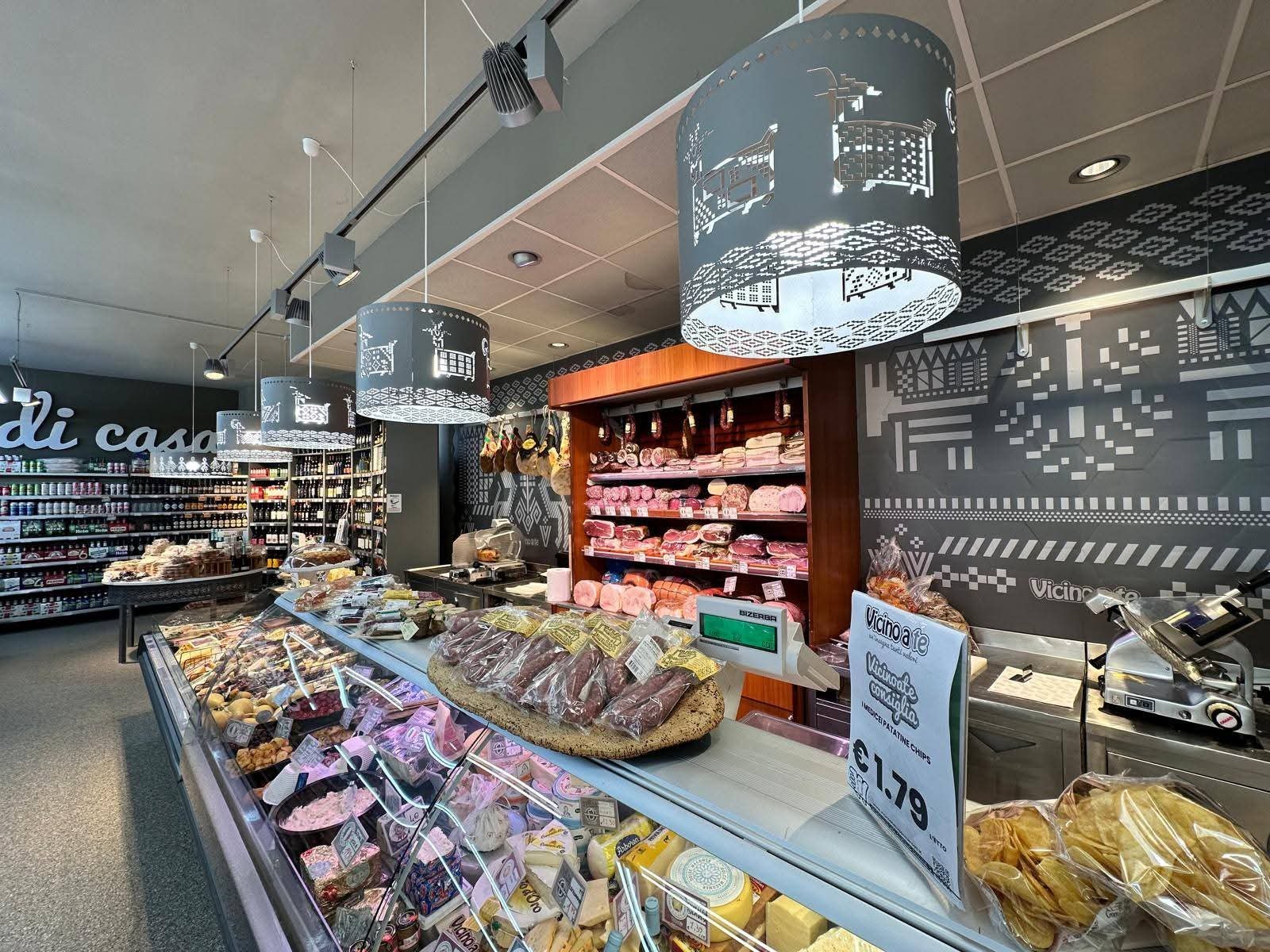 Banco salumi e formaggi Vicino a Te: prosciutti appesi, salsicce su tagliere, cartello patatine chips €1,79