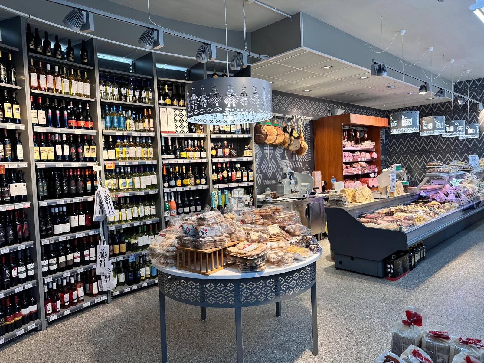 Interno Vicino a Te: scaffali di vini, tavolo di biscotti confezionati e banco salumi con forme di formaggio appese