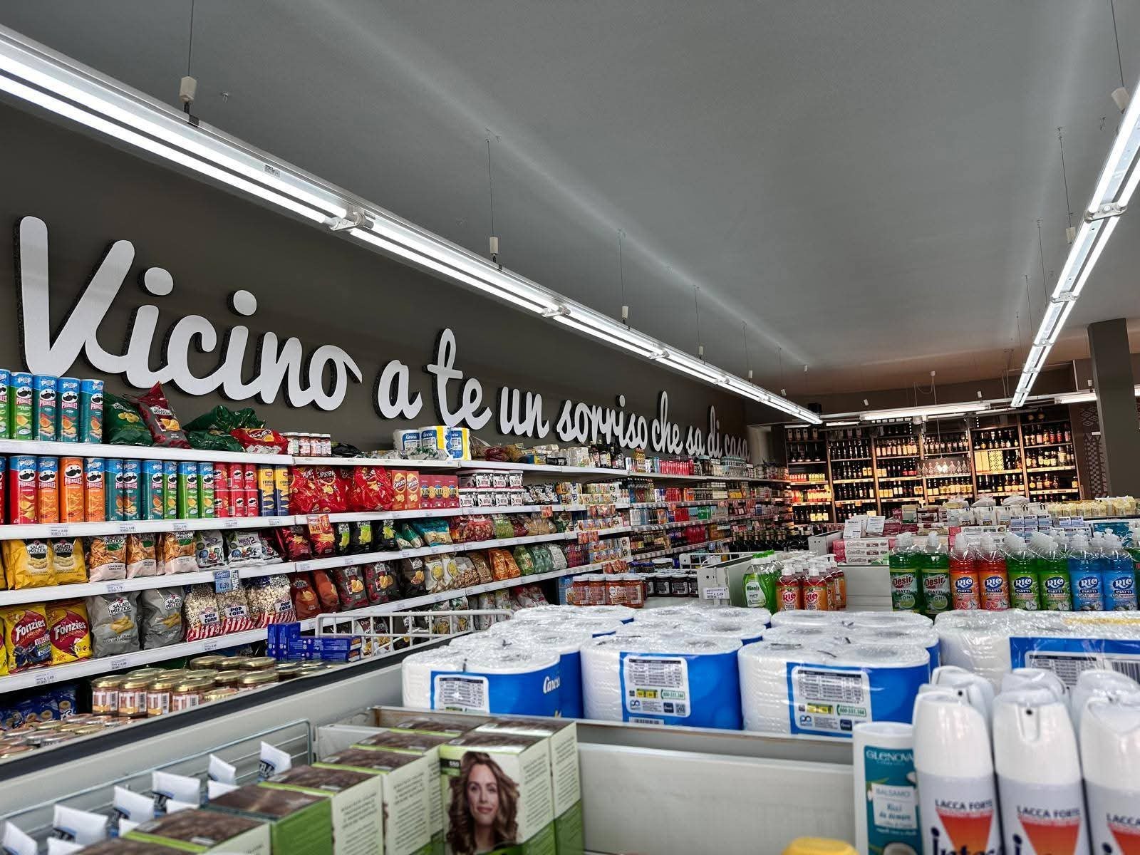 Corsia con snack, carta igienica e lacca sotto la scritta bianca «Vicino a te un sorriso che sa di casa», vini sul fondo