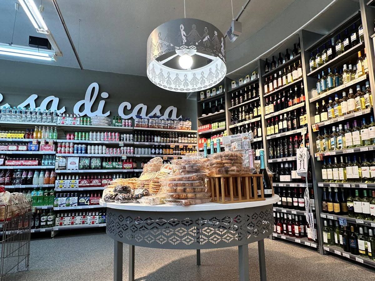 Tavolo con pane e biscotti confezionati sotto una lampada traforata, scaffali di vini e bibite, scritta murale «sa di casa